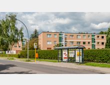 Bushaltestelle mit Wartehäuschen an einer Straße, im Hintergrund ein mehrstöckiges Wohngebäude mit beigefarbener Fassade und grünen Akzentelementen, umgeben von Hecken und vereinzelten Bäumen.