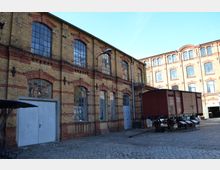 Fabrikgelände mit Backsteingebäuden im historischen Stil, gekennzeichnet durch große, bogenförmige Fenster und helle Klinkersteine mit roten Akzenten. Im Vordergrund steht ein brauner Güterwaggon auf einem Kopfsteinpflasterplatz.