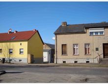 Zwei Wohnhäuser an einer ruhigen Straßenecke, eines mit gelber Fassade und rotem Ziegeldach, das andere mit grauer Putzfassade und dunklem Schieferdach. Im Vordergrund ist eine asphaltierte Straße mit einigen Rissen und Pfützen zu sehen.