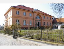 Zweistöckiges, sandfarbenes Gebäude mit roten Ziegeldach und bogenförmigem Eingang mit großem Rundbogenfenster. Vor dem Gebäude befindet sich ein Garten mit niedrigen Büschen, umgeben von einem Metallzaun; gepflasterter Weg im Vordergrund.