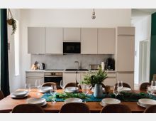 Moderne Küche mit weißen Schränken, grauer Rückwand und Edelstahlgeräten, inklusive Herd, Mikrowelle, Wasserkocher und Kaffeemaschine. Im Vordergrund steht ein gedeckter Esstisch mit Holztischplatte, weißen Tellern, Gläsern und einer Vase mit grünen Pflanzen.