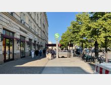 Straßenansicht mit einem historischen Gebäude auf der linken Seite, das Geschäfte wie Omega beherbergt, und einem grün-weißen S-Bahn-Schild, das den Eingang zur Station "Brandenburger Tor" markiert. Rechts sind Bäume, Fahrräder und eine belebte Straße mit Autos und Fußgängern zu sehen.