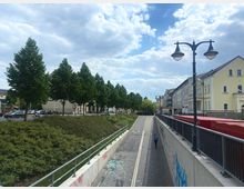 Straßenszene mit einer leicht abfallenden, asphaltierten Unterführung, flankiert von Betonmauern mit Graffiti. Auf der linken Seite stehen Bäume und geparkte Autos entlang einer erhöhten Straße, rechts befinden sich mehrstöckige Wohn- und Geschäftshäuser im klassischen Stil sowie Straßenlaternen.