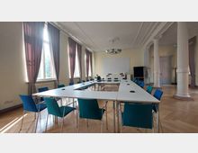 Heller Konferenzraum mit hohen Decken, großen Fenstern mit Vorhängen, weißen Wänden und dekorativen weißen Säulen. In der Mitte steht eine U-förmige Tischanordnung mit blauen Stühlen, ein Flipchart und eine Tafel sind im Hintergrund sichtbar.