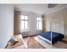 Schlafzimmer mit minimalistischer Einrichtung, bestehend aus einem weißen Himmelbett mit dunkelblauer Decke, einem Holzboden und einem Holztisch mit Schreibtischlampe im Vordergrund. Die Wände sind teilweise verputzt, zwei große Fenster mit beigen Vorhängen lassen Tageslicht herein.