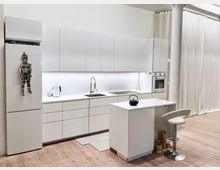 Moderne Küche mit weißen Hochglanzfronten, einer durchgehenden Arbeitsplatte, eingebautem Ofen, Spüle und Induktionskochfeld. Im Vordergrund steht ein weißer Küchentresen mit schwarzem Wasserkocher und passendem Barhocker; der Boden besteht aus hellem Holz, im Hintergrund hängen weiße Vorhänge.