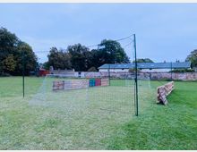 Grüne Rasenfläche mit einem kleinen improvisierten Fußballfeld, bestehend aus Netztoren und einer umgebenden Begrenzung aus Holzpaletten. Im Hintergrund stehen einige Bäume, eine Steinmauer sowie Gebäude mit flachen Dächern.