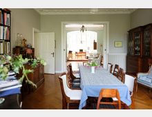 Esszimmer in einem Wohnhaus mit Holzboden, einem langen Tisch mit blauer Tischdecke und blühenden Tulpen. Im Hintergrund führt ein Durchgang in ein helles Wohnzimmer mit großen Fenstern, Schreibtisch und gepolsterten Möbeln.