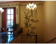 Treppenhaus eines historischen Gebäudes mit einer Wandmalerei im Zirkusstil, darunter Artisten und Tänzer. Im Hintergrund ein großes Fenster mit roten Vorhängen und ein Kronleuchter, der den Raum beleuchtet.
