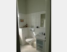 Kleines Badezimmer mit weißen Fliesen an Wänden und Boden. Zu sehen sind eine wandhängende Toilette, ein kleines Waschbecken mit Spiegelschrank darüber und einige Hygieneartikel auf einem Wandvorsprung.