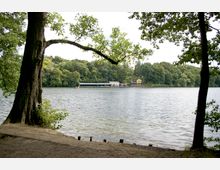 Uferansicht eines Sees, umgeben von dichtem Wald; im Hintergrund ein gelbes Haus und eine überdachte Anlegestelle, davor ein kleines Boot auf dem Wasser. Im Vordergrund stehen Bäume, deren Äste in die Szene ragen.