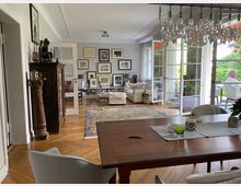 Wohnzimmer mit hellen Wänden, Holzboden und großen Glas-Schiebetüren, die zu einer Terrasse mit Gartenblick führen. Der Raum ist ausgestattet mit einer Holzanrichte, einem Esstisch, mehreren Sofas, einem Teppich sowie zahlreichen Bildern und Skulpturen, die die Wände und Möbel dekorieren.