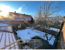 Ein Innenhof mit schneebedecktem Boden und Gebäuden aus roten Ziegelsteinen, die teils mit Rankgittern versehen sind. Im Vordergrund führen Metalltreppen hinunter, links und rechts säumen kahle Bäume und Hecken den Hof.