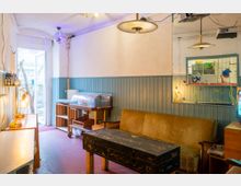 Ein kleiner, rustikaler Innenraum mit pastellblau gestrichenen Holzpaneelen und schlichter weißer Wand. Im Vordergrund ein alter brauner Ledersessel, ein abgenutzter Koffer als Tisch und eine Glasvitrine auf einem Holzgestell; seitlich ein offener Türbereich, durch den Bäume sichtbar sind.