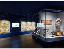 Innenansicht eines Museums mit Exponaten aus der japanischen Kultur. Zu sehen sind bemalte Paravents hinter Glasvitrinen, traditionelle Samurai-Rüstungen und Helme sowie weitere historische Artefakte, ausgestellt in einem abgedunkelten Raum mit blauer Beleuchtung.