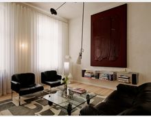 Modernes Wohnzimmer mit hohen Fenstern und bodenlangen weißen Vorhängen, hellen Holzdielen und einem gemusterten Teppich. Schwarze Ledersessel und Sofa gruppieren sich um einen gläsernen Couchtisch; an der Wand hängen ein großes dunkles Kunstbild über einem niedrigen Regal mit Büchern und es gibt eine schwenkbare Hängelampe.