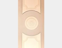 Dekorative Wandgestaltung mit weißen Stuckelementen: ein kreisförmiges Muster in der Mitte, eingerahmt von zwei rechteckigen Ornamenten mit geschwungenen Details. Die Wand hat einen hellbeigen Hintergrund.