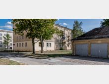 Wohnanlage mit hellen mehrstöckigen Gebäuden und Balkonen, umgeben von Bäumen und Rasenflächen; rechts eine kleine Garage mit geschlossenen Toren. Der Himmel ist klar und blau.
