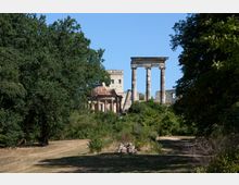 Ruinen eines historischen Gebäudes mit Säulen und einem Burgähnlichen Teil im Hintergrund, umgeben von dichten Bäumen und einer Wiese im Vordergrund. Der Himmel ist klar und blau.