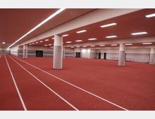 Großer, moderner Innenraum mit rotem Gummiboden, der eine Laufbahn und eine freie Fläche umfasst, unterstützt von weißen, runden Säulen; helle rechteckige Deckenleuchten beleuchten den Raum, umgeben von grauen Wänden mit Türen und einer Rampe.