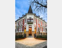 Ein historisches Gebäude mit spitzem Turmdach, weißer und roter Fassade sowie dekorativen Fensterelementen. Vor dem Eingang führt ein gepflasterter Weg durch ein umzäuntes Grundstück, umgeben von kahlen Bäumen und blauem Himmel.