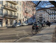 Straßenszene in einer Wohngegend mit historischen Gebäuden, die helle Fassaden und Balkone haben. Auf der gepflasterten Straße sind Autos geparkt, ein Fahrrad steht am Gehweg, und im Hintergrund läuft eine Person auf dem Bürgersteig.