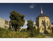 Eine Backsteinkirche mit achteckigem Grundriss und hohem Turm in neugotischem Stil, umgeben von Bäumen und einer Wiese. Links im Bild befinden sich Wohnhäuser, und ein Metallzaun trennt die Grünfläche vom Gehweg.