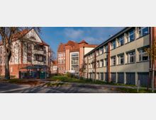 Außenansicht eines Schulgebäudekomplexes mit roten Klinkerfassaden und großen Fenstern. Links ein historisches Gebäude mit Fachwerkelementen, rechts moderne Schulgebäude, umgeben von Grünflächen und einem Gehweg.