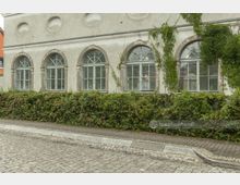 Fassade eines historischen Gebäudes mit mehreren großen Rundbogenfenstern, teilweise mit Efeu bewachsen. Vor dem Gebäude verläuft ein schmaler Gehweg, eingefasst von einer Hecke; davor eine gepflasterte Straße.