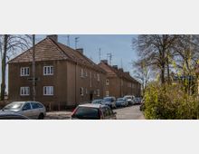 Straßenszene mit mehreren zweigeschossigen Wohnhäusern aus braunem Putz und roten Satteldächern, daneben geparkte Autos. Im Vordergrund stehen Bäume und Büsche, im Hintergrund ein Straßenschild.