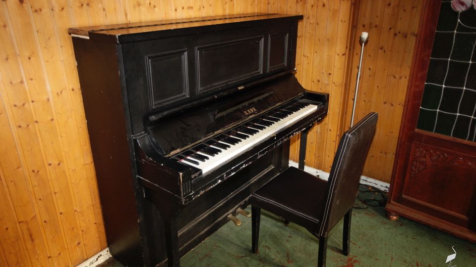 Schwarz lackiertes Klavier mit Hocker in einem Raum mit holzvertäfelten Wänden und grünem Teppichboden. Rechts steht ein dunkler Holzschrank mit Glaseinsatz neben einer schlanken Stehlampe.