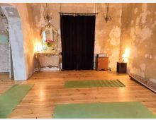 Ein Raum mit hölzernem Boden, mehreren grünen Yogamatten, einer cremefarbenen Wand mit Patina-Spuren, einem goldenen Spiegel auf einem kunstvoll verzierten Tisch und einem schwarzen Vorhang. Beleuchtung erfolgt durch Tischlampen und Wandkerzenhalter.