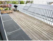 Holzterrasse mit Geländer, ausgestattet mit einem Holztisch, Stühlen, einem Grill und einem bepflanzten Blumenkasten; im Hintergrund sind umliegende Gebäude und Bäume zu sehen.