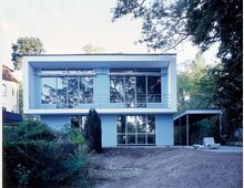 Modernes zweistöckiges Gebäude mit weißer Fassade und großen Glasfenstern, umgeben von Bäumen. Im Vordergrund befindet sich ein Gartenbereich mit Büschen, ein schmales Vordach und minimalistische Gartenmöbel.