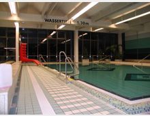 Ein Hallenbad mit gefliesten Böden, einem rechteckigen Schwimmbecken und einer roten Rutsche. Im Hintergrund sind große Fenster und Pflanzen zu sehen, darüber der Hinweis auf die Wassertiefe von 1,30 m.