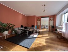 Wohnzimmer mit Holzboden, einer terrakottafarbenen Wand und einer grauen Eckcouch. Ein großer schwarzer Teppich bedeckt teilweise den Boden, daneben stehen Pflanzen und moderne Möbel. Im Hintergrund führt eine offene Tür in einen weiteren Raum, wobei an den Seiten zwei große, bemalte Holzfiguren stehen.