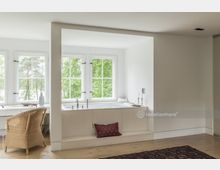 Helles Badezimmer mit freistehender Badewanne vor einer Fensterfront mit weißen Sprossenfenstern und Blick ins Grüne. Neben der Badewanne befindet sich ein kleiner Schreibtisch mit einem Stuhl aus hellem Rattan.