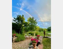 Steingepflasterte Terrasse mit Gartenmöbeln und Blumentöpfen, umgeben von grünen Bäumen und Rasen; im Hintergrund sind zwei Regenbögen vor einem blauen Himmel zu sehen.