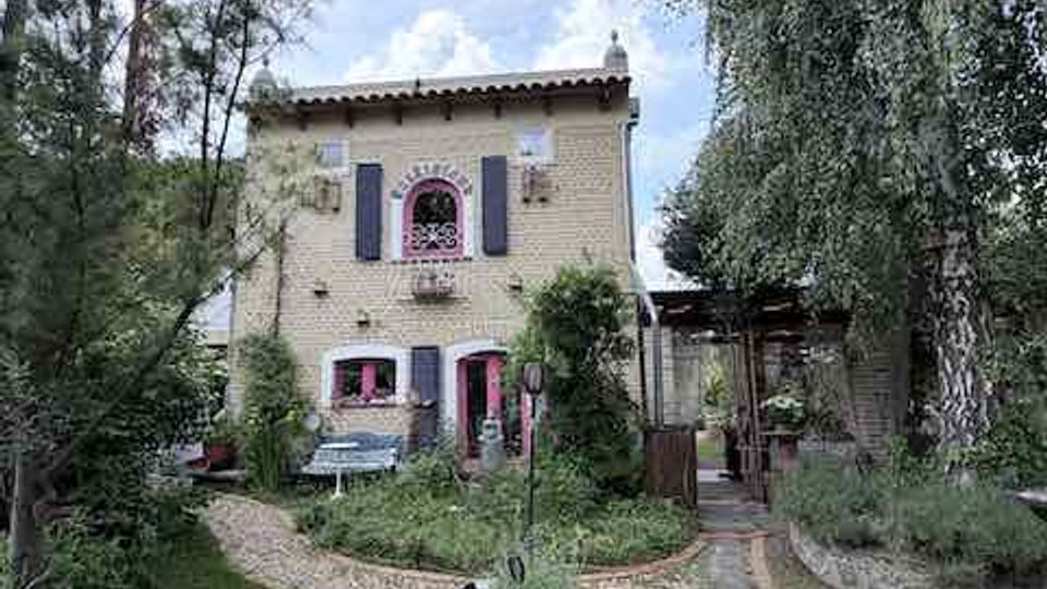 Zweistöckiges Haus aus hellem Ziegelstein mit violetten Fensterläden und roter Eingangstür, davor ein Garten mit Rasen, Kieswegen und Pflanzkübeln. Hohe Bäume und Sträucher rahmen das Gebäude ein, rechts führt ein überdachter Durchgang zu einem Nebengebäude.