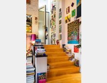 Ein Wohnraum mit hölzernen Treppenstufen, gesäumt von gestapelten Büchern. Wände sind mit farbenfrohen Gemälden dekoriert, und Regale voller Bücher erstrecken sich bis zur Decke.