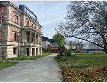 Historisches Gebäude mit neoklassizistischen Säulen und rotem Ziegelmuster, umgeben von einem gepflegten Rasen und vereinzelten Bäumen; im Hintergrund moderne Gebäude und ein kleiner Parkplatz. Ein gepflasterter Weg führt entlang des Gebäudes durch die Grünanlage.