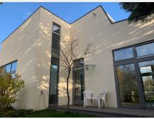 Modernes Wohnhaus mit heller Fassade und großen, rechteckigen Fensterflächen. Im Vordergrund eine kleine Terrasse mit zwei weißen Kunststoffstühlen, ein kahler Baum und ein Wasserschlauch an der Wand; umgeben von einer Grünfläche mit Sträuchern.