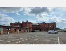 Großer, gepflasterter Parkplatz vor mehreren roten Backsteingebäuden im industriellen Stil, darunter ein altes Gebäude mit Runddach und ein mehrstöckiger Komplex mit Bogentüren und vielen kleinen Fenstern. Im Vordergrund stehen geparkte Autos, und der Himmel ist teilweise bewölkt.