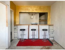 Kleine Bar mit drei schwarzen Barhockern vor einer Theke aus Holz und Stein, die durch eine mit goldenem Putz verkleidete Rückwand hervorgehoben wird. Im Hintergrund befindet sich ein Regal sowie eine schlichte Deckenleuchte, der Boden besteht aus Beton mit einem roten Teppich vor der Theke.