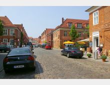 Straßenszene in einer Altstadt mit Kopfsteinpflaster, gesäumt von niedrigen Backsteingebäuden mit roten Dächern. Auf beiden Seiten sind geparkte Autos, Straßencafés mit orangefarbenen Sonnenschirmen und Fußgänger erkennbar.