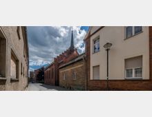 Schmale gepflasterte Gasse mit historischen Gebäuden aus Backstein und Putzfassaden, rechts die Seitenwand einer Kirche mit spitzem Glockenturm und kleinen Spitzbogenfenstern. Im Hintergrund ein weiteres historisches Gebäude und ein wolkiger Himmel.