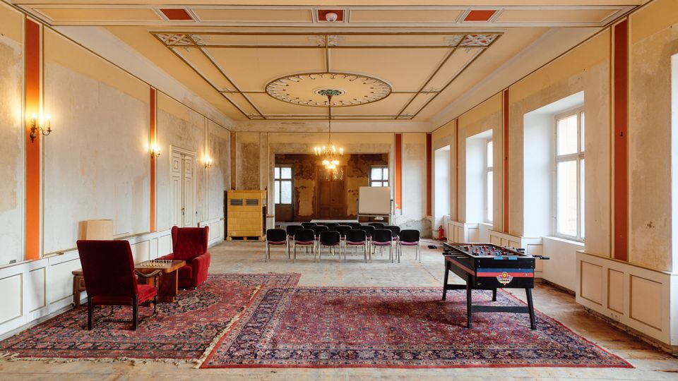 Großer, rechteckiger Saal in einem älteren Gebäude mit hohen Decken, Stuckornamenten und Kronleuchter sowie hohen Fenstern entlang einer Wand. Im Raum stehen Sitzreihen, Teppiche, zwei Sessel mit Beistelltisch und ein Tischkicker; die Wände sind teilweise abgeblättert und renovierungsbedürftig.