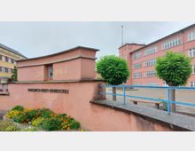 Außenansicht der Friedrich-Ebert-Grundschule mit einem rosa Gebäude im Hintergrund, zahlreichen rechteckigen Fenstern und einer Uhr an der Fassade. Im Vordergrund eine rosa Mauer mit einer Gedenktafel, darauf ein Blumenbeet und präzise geschnittene kleine Bäume.