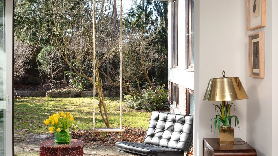 Modern living room with white walls and a concrete ceiling, featuring a large sliding glass door that opens to a backyard garden with trees and a hanging swing. Inside are a black tufted lounge chair, a small red table with yellow flowers, and a wooden side cabinet with a brass lamp and framed artwork.