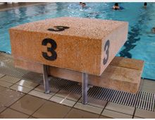 Startblock Nummer 3 in einem Hallenbad, platziert an einem gefliesten Beckenrand mit mehreren Personen, die im Wasser schwimmen.